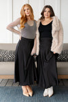 Two women wearing black maxi skirts, one with a cascading hem and the other with a flat front, posing on a sofa with a decorative faux fur coat on top.
